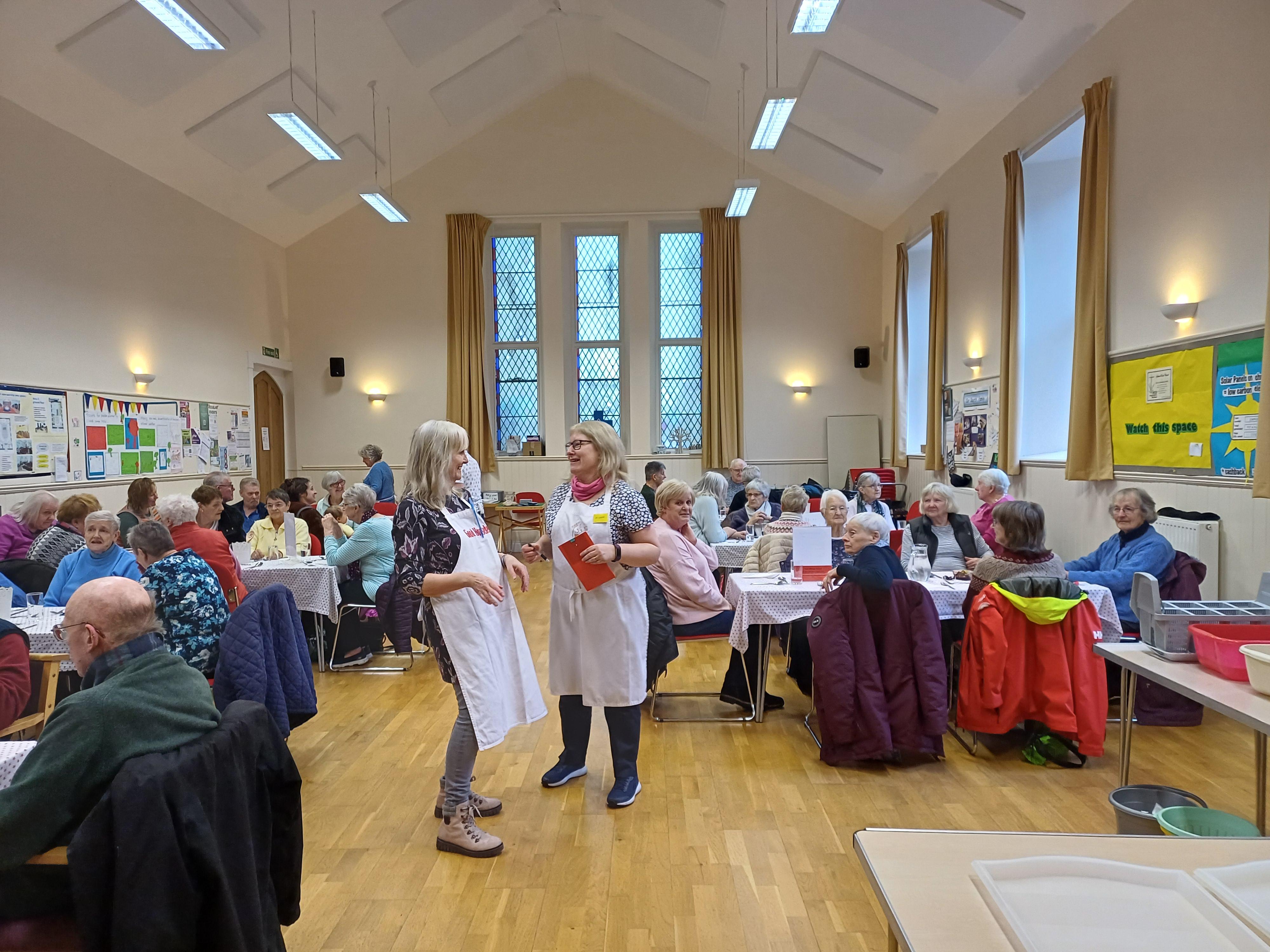 Two women in a church hall laugh. They are surrounded by many people eating while sat down at tables; a community hub.