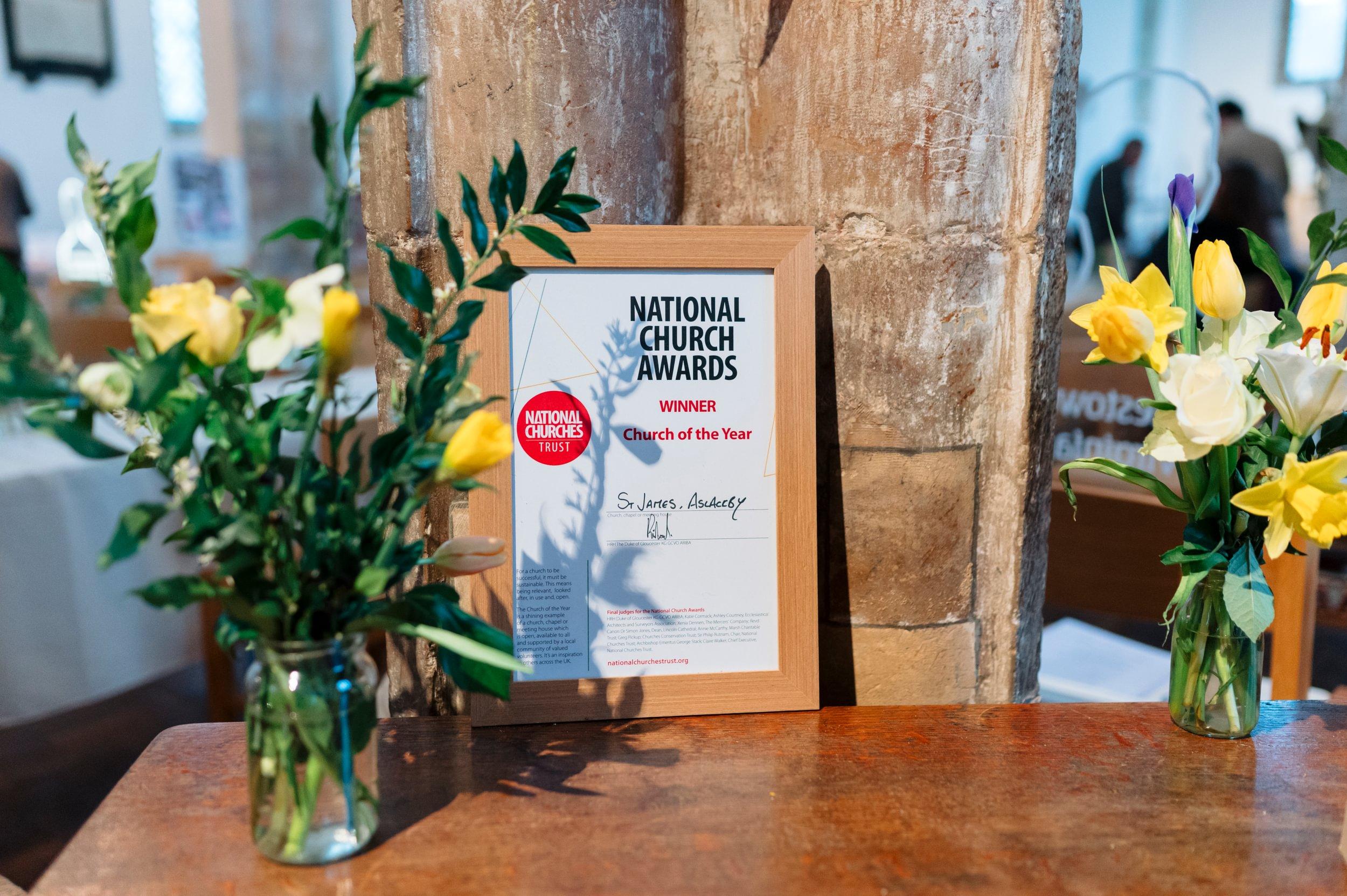 A wooden table with a certificate displayed, which congratulates St James the Great as the Church of the Year 2024
