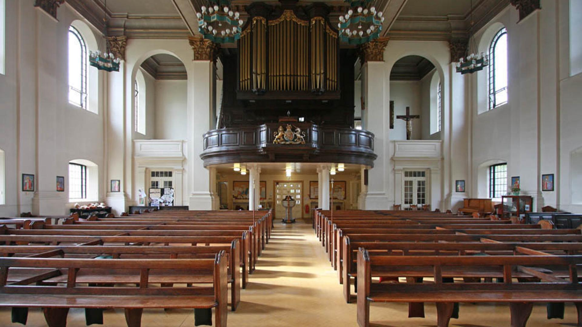 The interior of All Saints in Poplar, taken from the altar looking over rows of benches to the back of the church where an organ is.