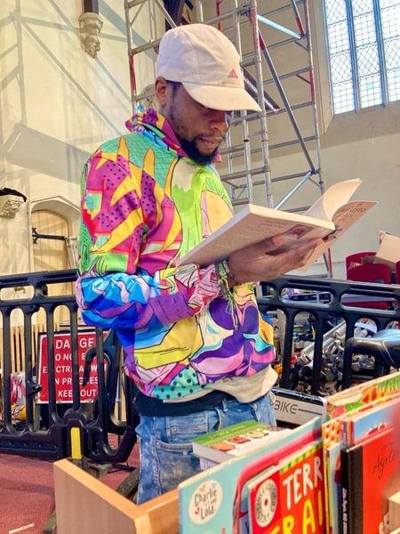 A man wearing a multi-coloured jacket and baseball cap reads a book inside a church