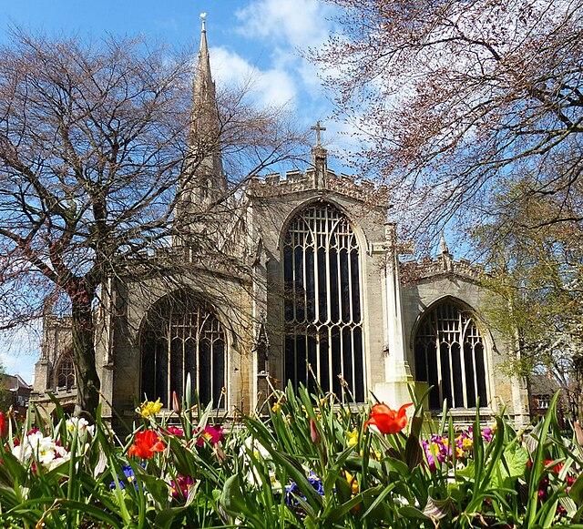 A large church - Newark St Mary Magdalene in Newark - with large windows. There are bare trees and colourful flowers in the foreground and a blue sky with few clouds.