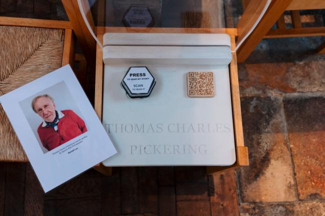 A chair with a large button on it; this is part of the audio trail at the church. A piece of laminated paper is next to this, which shows a photo of David on it.