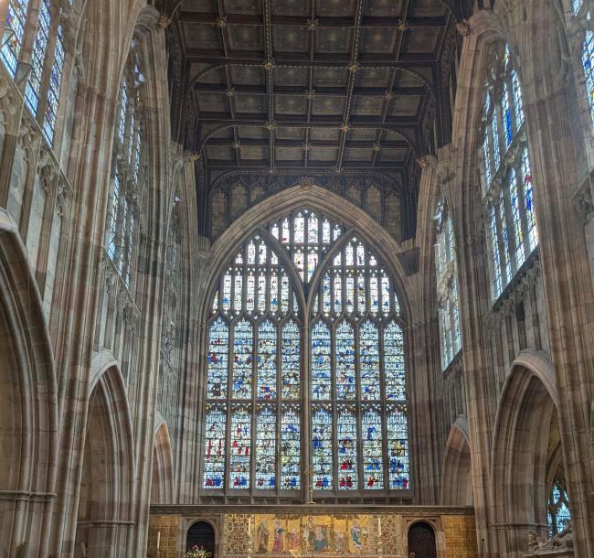 A photo of some of the beautiful stained glass at Great Malvern Priory. You can see one large arched window and several smaller windows on the left and right hand side.