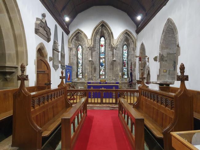 The stunning interior of Medomsley St Mary Magdalene in County Durham. Two wooden pews facing each other sit infront of an altar. There is grey stone around the stained glass windows above the altar.