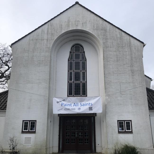 A grubby white church, with a banner outside the front reading 'Paint All Saints'