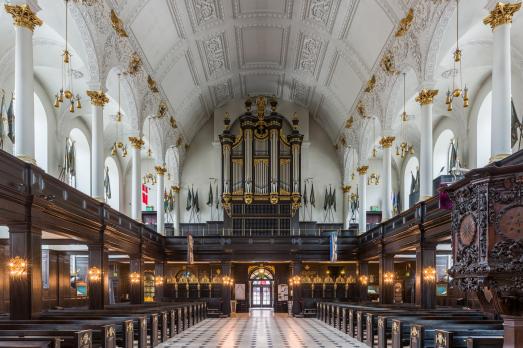 Interior of St Clement Danes church. A barrel-vaulted moulded ceiling painted white with gold details. The pews and panneling on the walls are in very dark wood. The organ sits at the far end of the church.