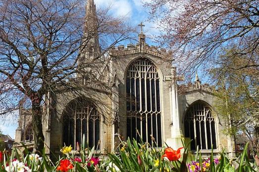 A large church - Newark St Mary Magdalene in Newark - with large windows. There are bare trees and colourful flowers in the foreground and a blue sky with few clouds.